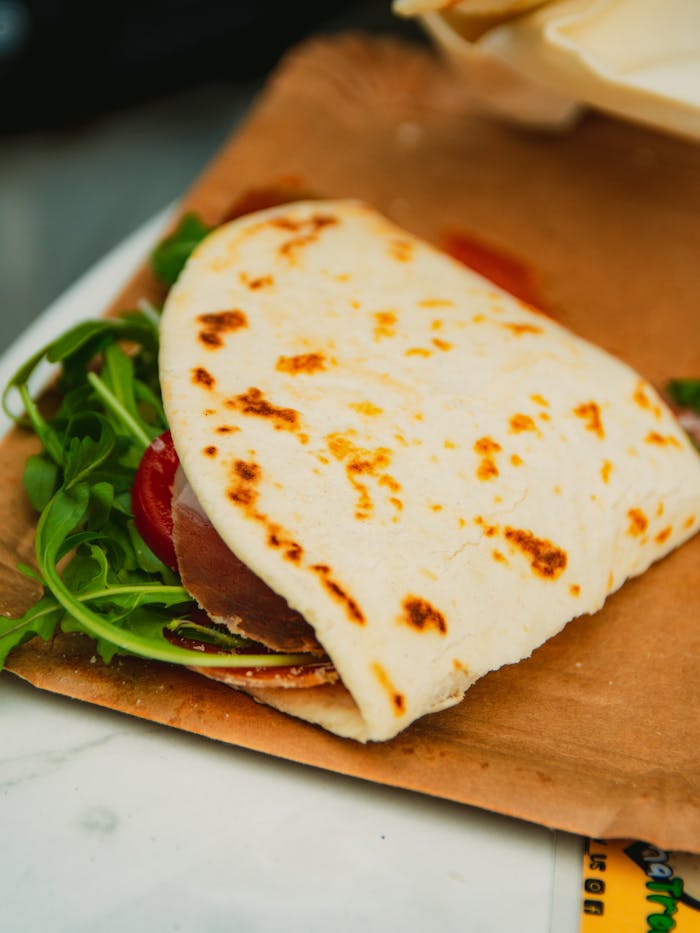 Tempting piadina romagnola filled with arugula, prosciutto, and tomato on a paper plate.